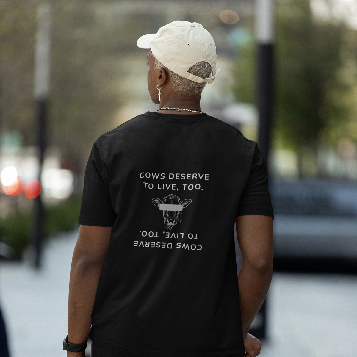 Mann von hinten mit einer beigen Cap, einem silbernen Ohrring und einem schwarzen T-Shirt mit folgendem Aufdruck auf dem Rücken: "Cows deserve to live, too." sowie einer Kuh mit einem grauen Balken vor den Augen.