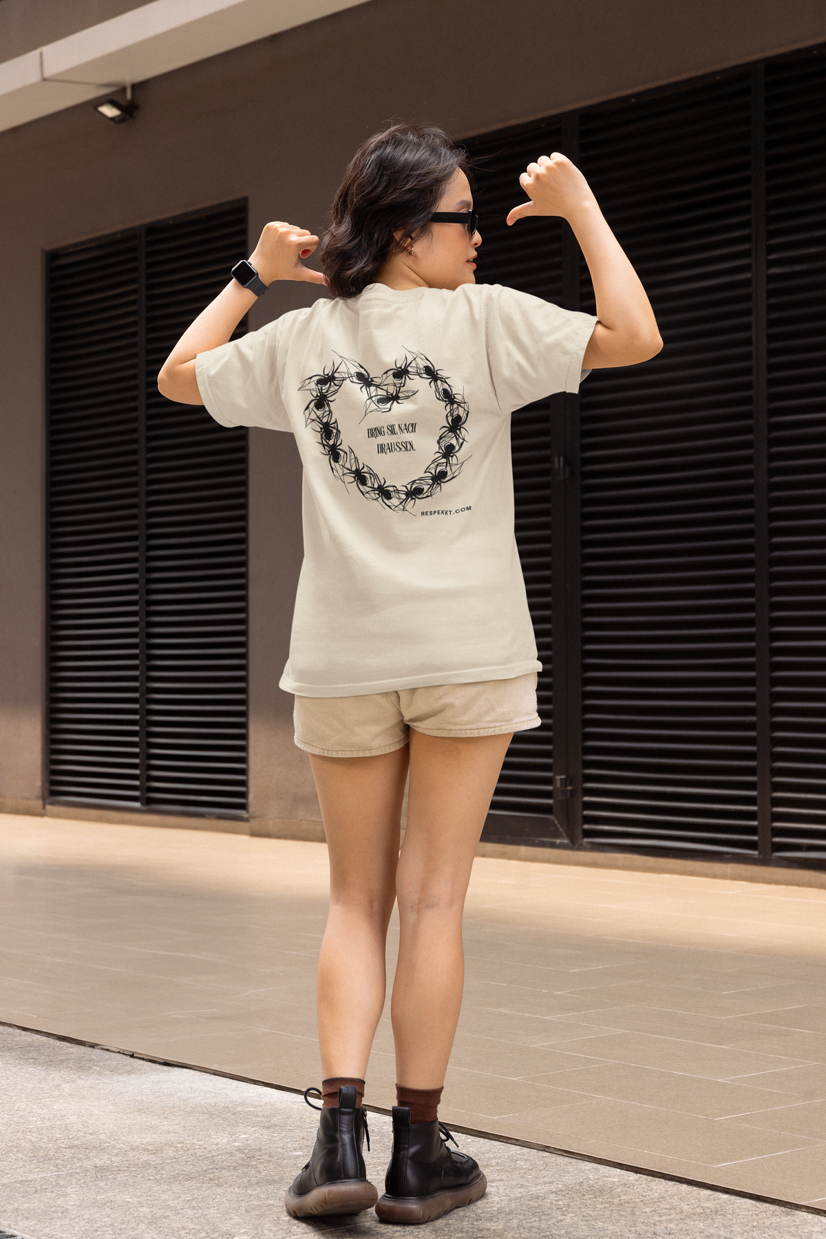 Ganzkörperaufnahme einer Frau, welche  beige Hotpants trägt und ein weißes T-Shirt mit dem Aufdruck: "Bring sie nach draußen." und einem Herz geformt aus Spinnen um den Schriftzug. Die Frau zeigt mit ihren Daumen auf die Rückseite des T-Shirts.