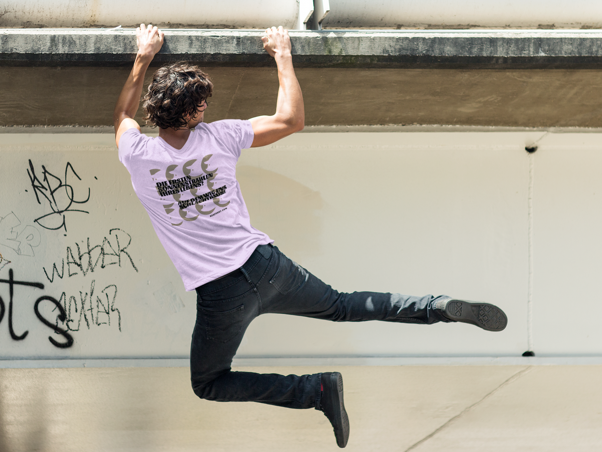 Person, die sich mit seinen Händen an einem Mauervorsprung festhält. Auf dem lilafarbenen T-Shirt sind Schweineköpfe aufgedruckt und der Schriftzug lautet: "Die ersten Sonnenstrahlen ihres Lebens -auf dem Weg ins Schlachthaus."