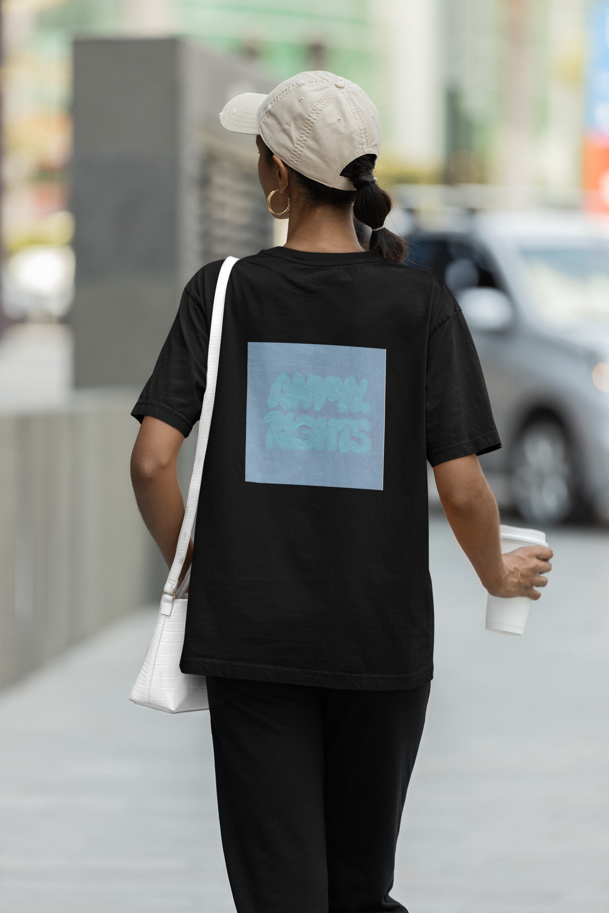 Frau von Hinten mit schwarzem T-Shirt mit dem türkisen Aufdruck: "Animal Rights". Sie trägt eine beige Cap, Handtasche, sowie einen Kaffeebecher in ihrer Hand.