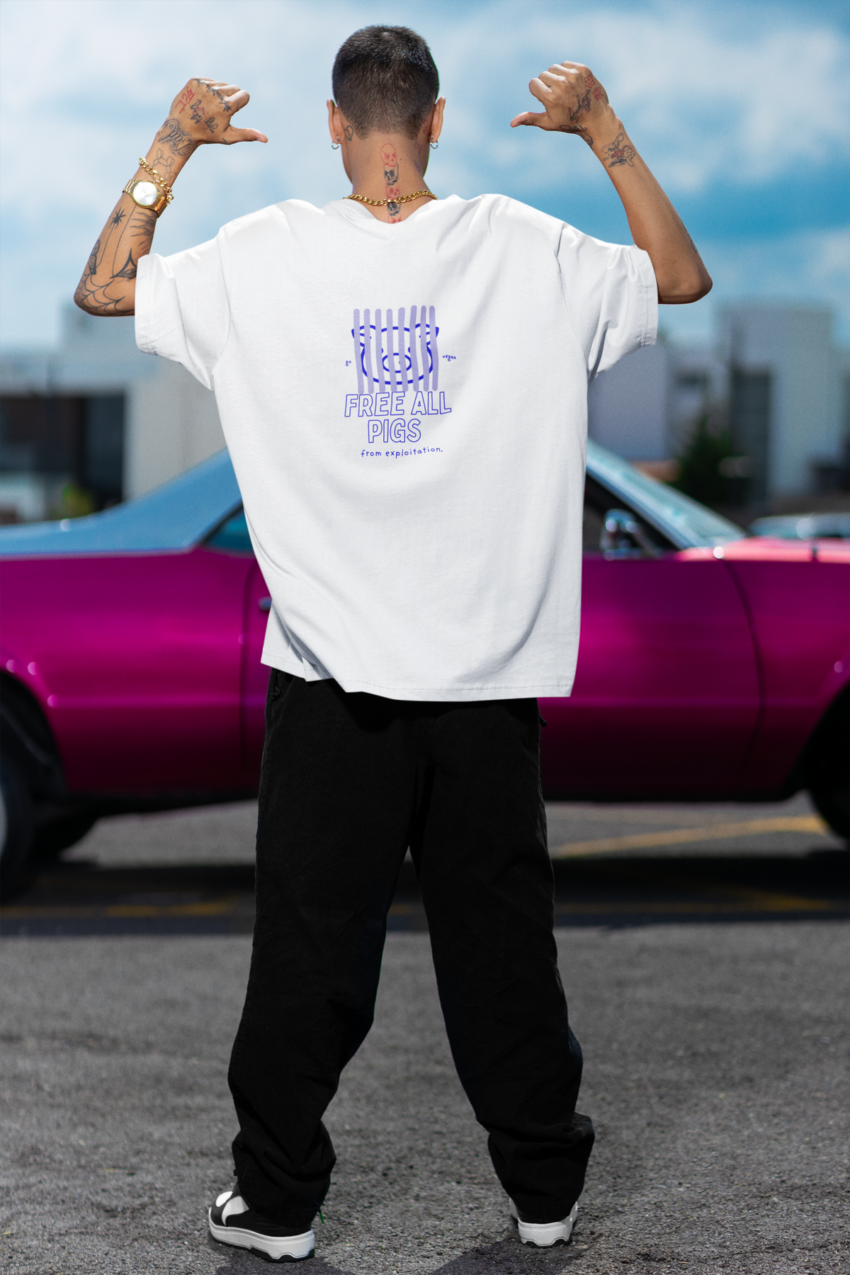 Tättowierter Mann der ein weißes T-Shirt trägt mit dem Aufdruck auf dem Rücken: "Free all pigs from exploitation." Die Schrift ist in blauer Farbe und es ist ein Schwein hinter Gitterstäben abgebildet. Im Hintergrund befindet sich ein Auto.
